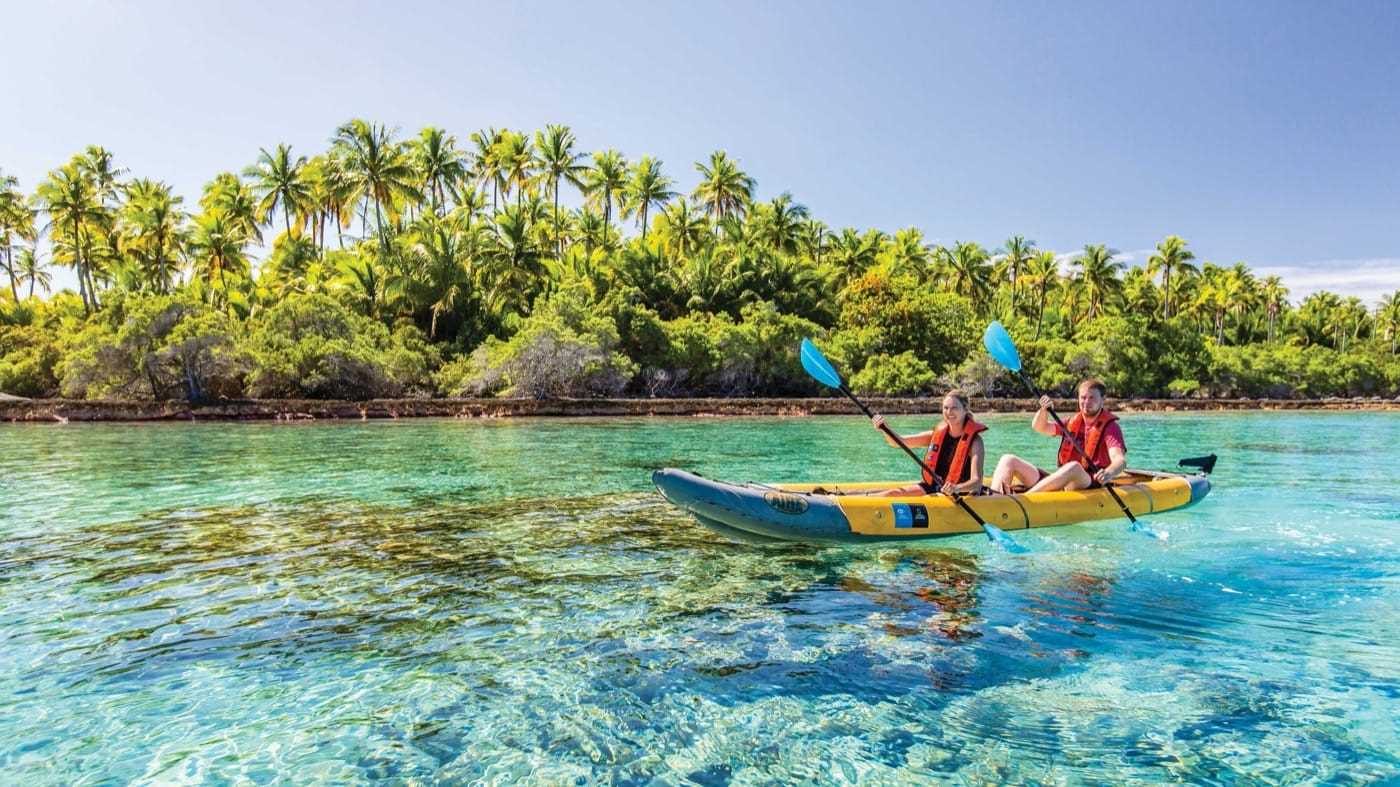 Two people rowing a boat through crystal-clear water close by a verdant shoreline