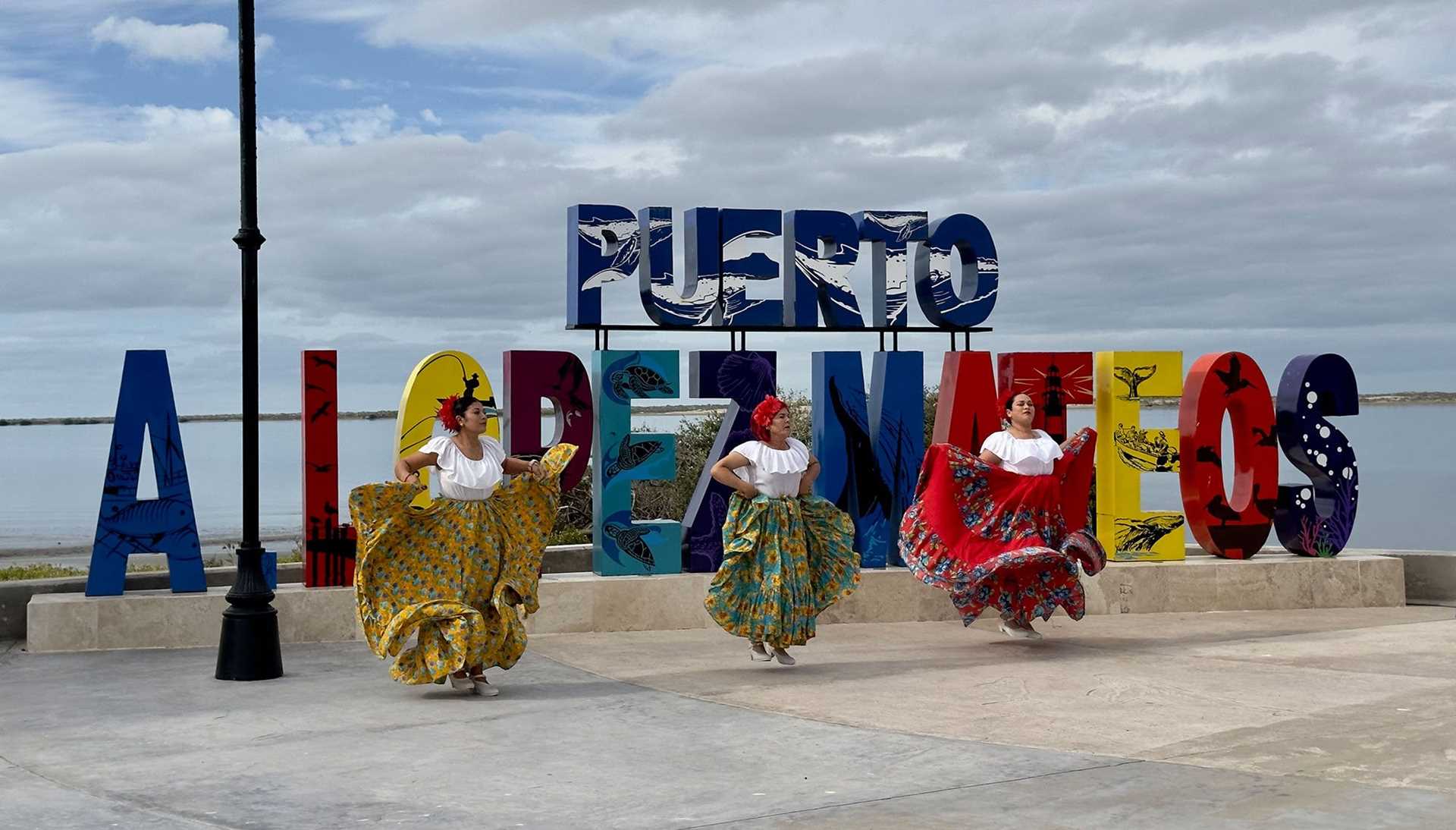 three dancers in colorful costumes in front of a colorful sign that says Puerto a Lopez Mateos