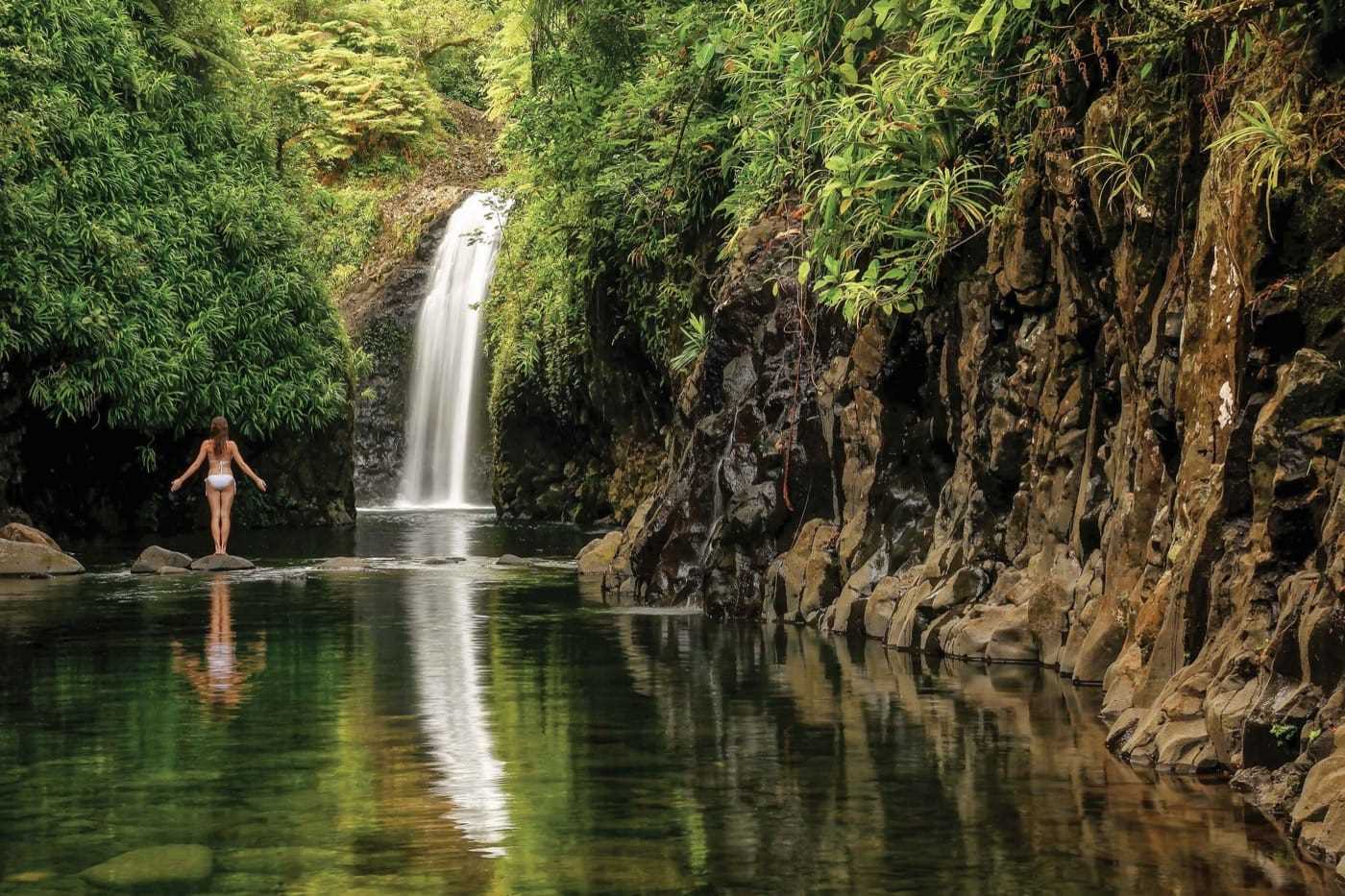 Lush greenery, a small waterfall and rocky cliffs surround a peaceful pool of water; where a lone woman practices yoga