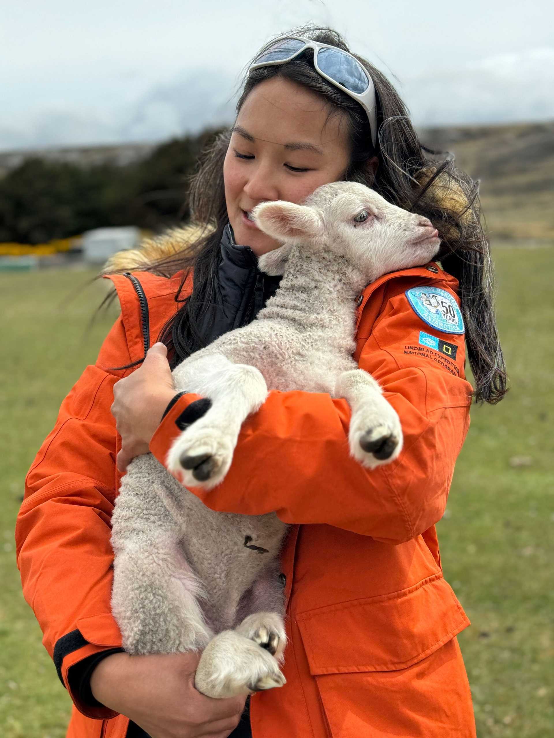 a woman in an orange parka holds a baby lamb