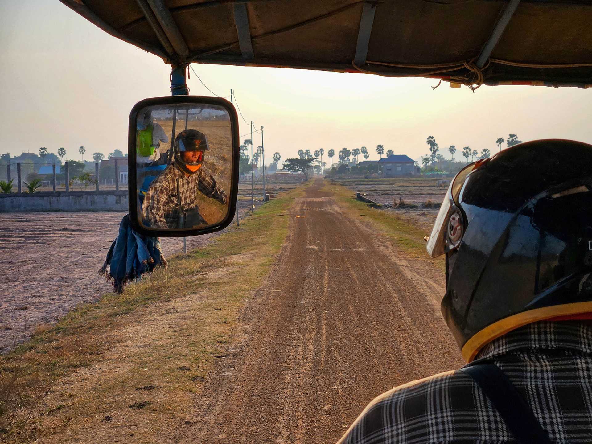 a tuk-tuk driver visible in the rear view mirror of his vehicle