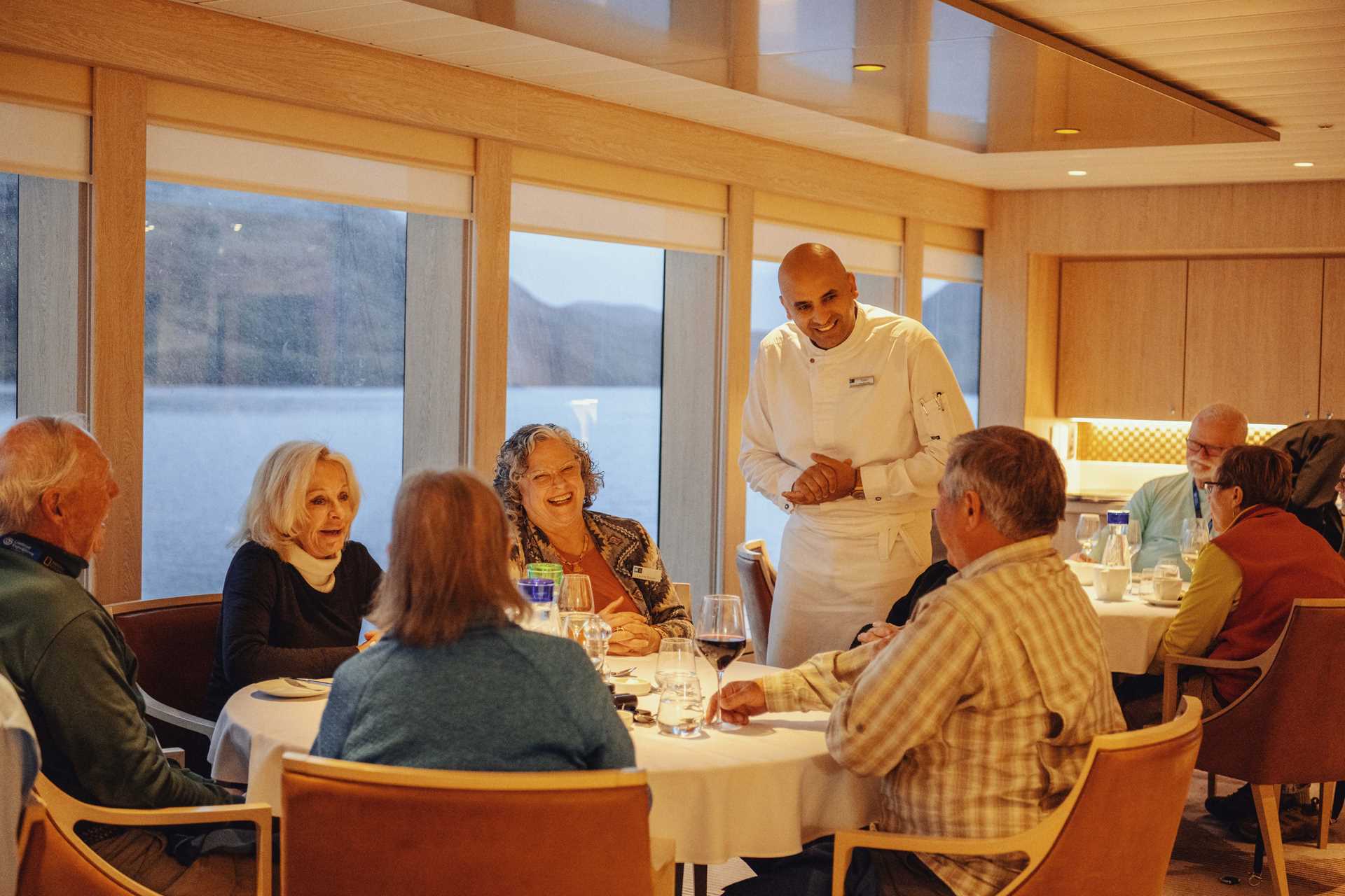 Five guests, three female and two male, eat at a dinner table while the chefs explain their meal.