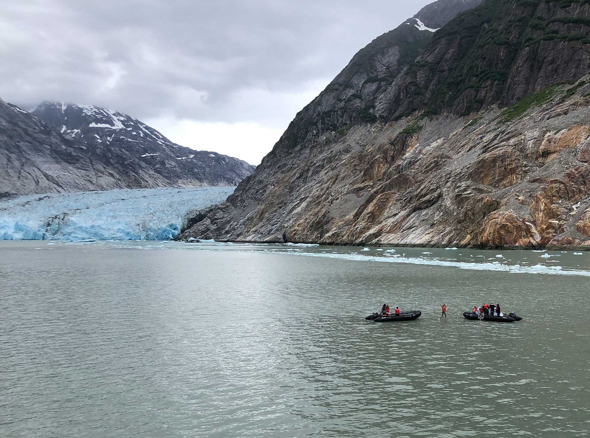 a person in a red jacket jumps off a zodiac in front of a large glacier